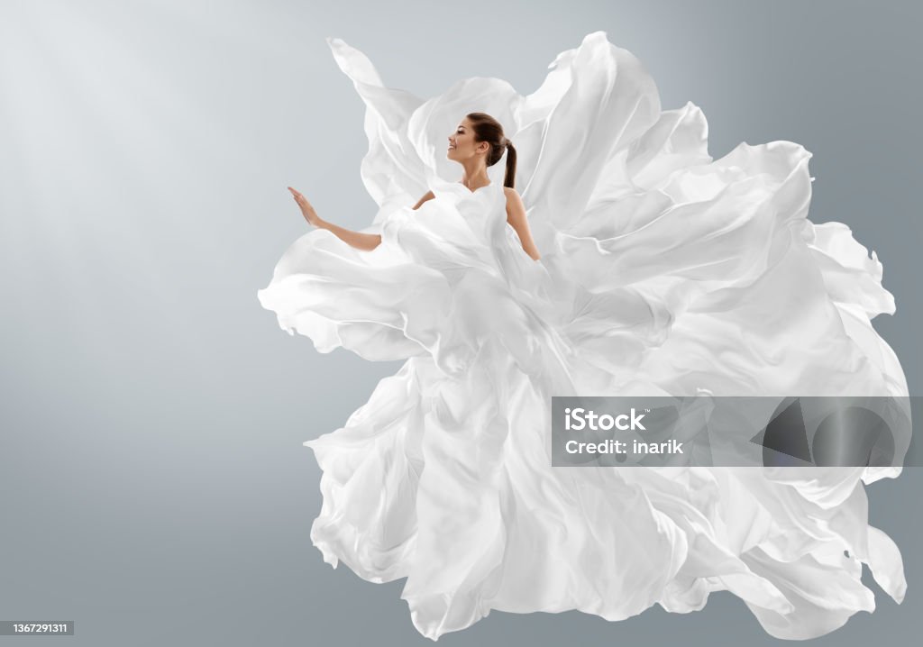 Fashion Model in Creative Pure White Dress as Cloud. Woman in Long Silk Gown with Chiffon Fabric flying on Wind over Light Gray Background. Art Fantasy dancing Girl Fashion Model in Creative Pure White Dress as Cloud. Woman in Long Silk fluttering weightless Gown with Chiffon Fabric flying on Wind . Art Fantasy dancing in Air Girl over Light Gray Studio Background White Color Stock Photo