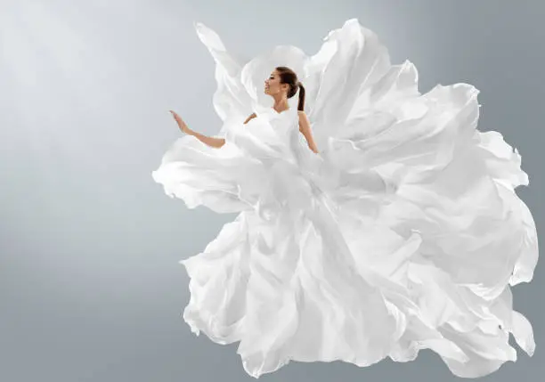 Fashion Model in Creative Pure White Dress as Cloud. Woman in Long Silk fluttering weightless Gown with Chiffon Fabric flying on Wind . Art Fantasy dancing in Air Girl over Light Gray Studio Background