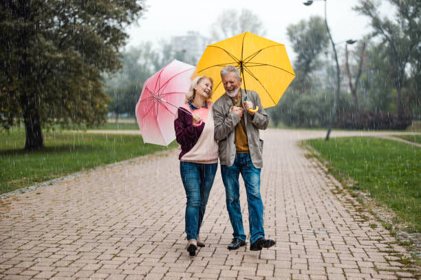 cheerful senior couple talking while walking on rainy day in nature. - umbrella senior adult couple autumn imagens e fotografias de stock