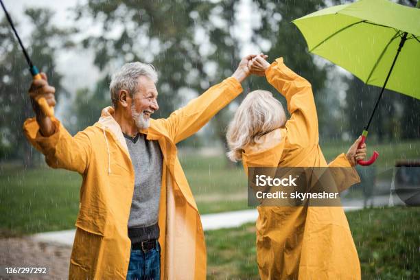 Carefree Senior Couple In Raincoats Dancing On A Rainy Day Stock Photo - Download Image Now