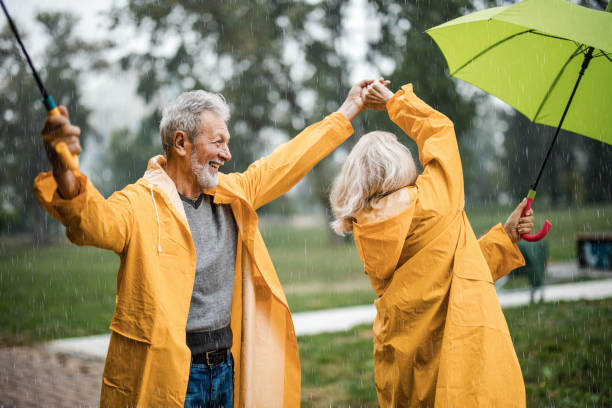 carefree senior couple in raincoats dancing on a rainy day. - umbrella senior adult couple autumn imagens e fotografias de stock