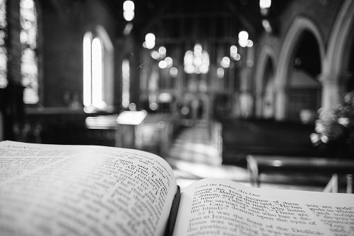Monochrome image depicting a close up wide angle view of the pages of an open bible on the altar of an Anglican church. Focus is on the bible in the foreground, while the background consists of the defocused interior of the church. Wooden pews recede into the distance and the lights illuminating the church appear as out of focus balls of light. The church has a warm, welcoming orange glow. Lots of room for copy space.