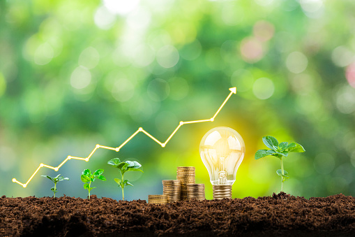 Close up view of a financial growth graph, coin stacks, plant pods and a glowing light bulb on soil shot against defocused lush foliage green background. Focus on foreground
