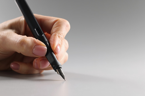 Female fingers hold a black fountain pen on a gray background, close-up. Correspondence concept, copywriting