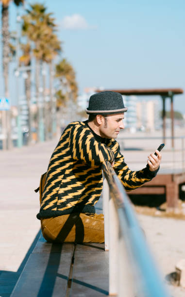 closeup of a mature man, wearing colorful modern casual clothes, watching his smartphone, sitting outdoors next to the sand of a urban beach
