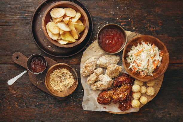 Asian-style barbecue chicken drumsticks and wings with coleslaw and potato chips. Flat lay top-down composition on dark wooden background.