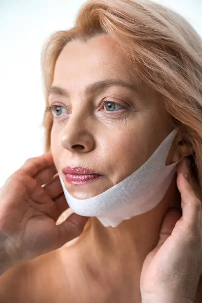 Female person looking away with calm and serious expression while applying mask from wrinkles on her chin. Self care concept