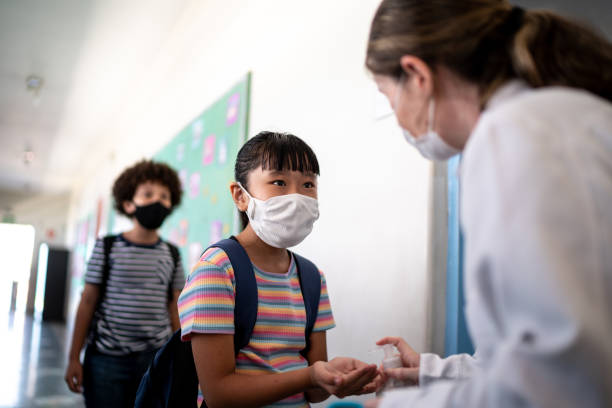 istock Teacher applying hand sanitizer on student's hand before entering classroom - using a face mask 1367325003