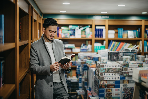 handsome young man reading a book at the library or bookstore leaning on the shelf copyspace interesting lifestyle hobby education learning studying project information youth.