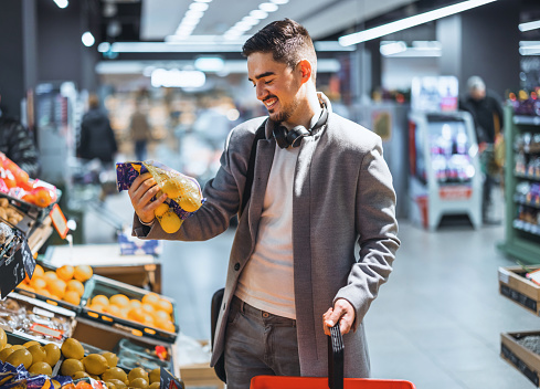 young asian man chooses and picks in eco bag apple fruit or vegetables in the supermarket. male customer standing a grocery store near the counter buys and throws in a reusable package in market