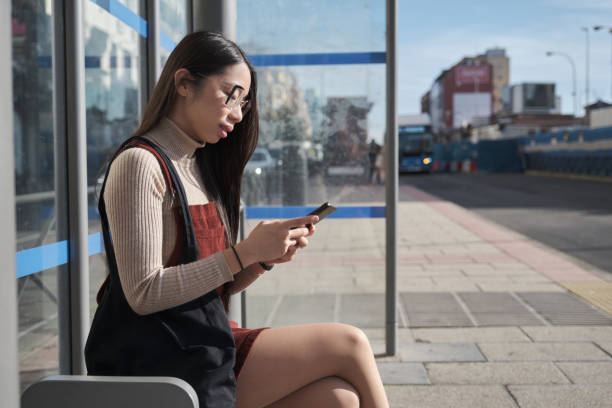 istock Young asian woman using her smartphone while waiting for the bus at bus stop. 1367331180