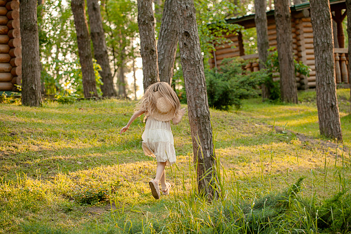 Rear view of preteen girl with long blonde hairs in light dress and straw hat running among tall trees in green summer city park