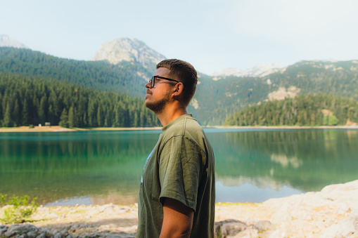 Happy man in eyeglasses hiking around the picturesque Black Lake with view of the mountains and pine forest in Durmitor National park