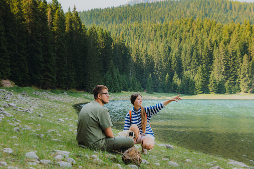 Happy heterosexual couple and their dog - pug breed sitting by the picturesque Black Lake with view of the mountains and pine forest in Durmitor National park