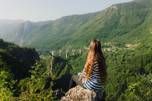 Rear view of woman with long hair in striped shirt sitting on the rock with viewpoint of the Tara canyon and the huge bridge in Durmitor National park