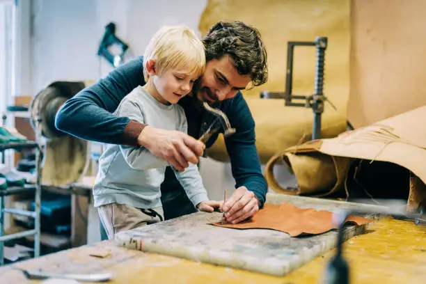 Smiling father is teaching his son making luxury leather belt in the workshop.