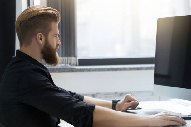istock Side view of a young bearded man sitting at his desk 1367343349