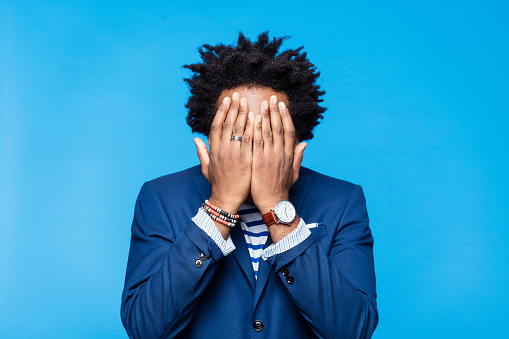 Headshot of afro american man wearing blue jacket, striped t-shirt, covering face with hands. Studio shot on blue background. Portrait of designer.