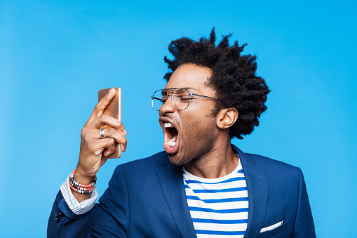 Headshot of upset afro american man wearing navy blue jacket, striped t-shirt and eyeglasses, shouting into smart phone. Studio shot on blue background. Portrait of designer.