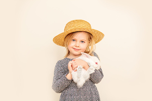 Girl child is cute and happy with a white rabbit in her hands against a white wall in a braided hat