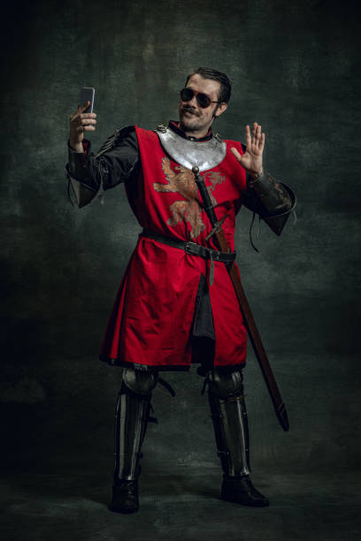 istock Smiling young man in image of medieval warrior or knight with dirty wounded face using phone isolated over dark background. Comparison of eras 1367355121