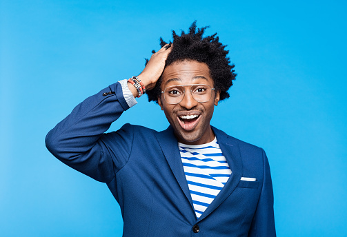 Afro american man wearing navy blue jacket, striped t-shirt and eyeglasses looking at camera with mouth open and hand in hair. Studio shot on blue background. Portrait of designer.