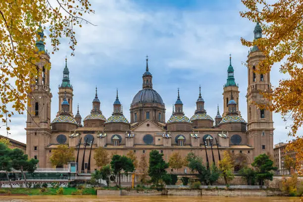 Spanish Cathedral Basilica Nuestra Senora del Pilar among golden autumn trees on the embankment of the river Ebro with yellow water in Zaragoza. Beautiful architectural background with copy space
