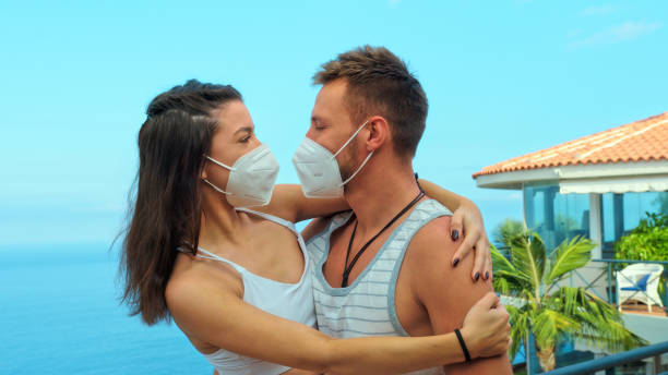 Couple on a patio with sea view, hugging in protective masks (type FFP2). Love in the times of pandemic Sea panorama seen from a patio. Vacations memories vacation rental mask stock pictures, royalty-free photos & images