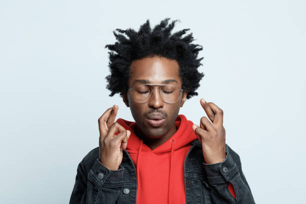 Headshot of man crossing fingers Cheerful afro american young man wearing red hoodie, black jeans jacket and glasses, crossing fingers with eyes closed. Studio shot on grey background. fingers crossed stock pictures, royalty-free photos & images