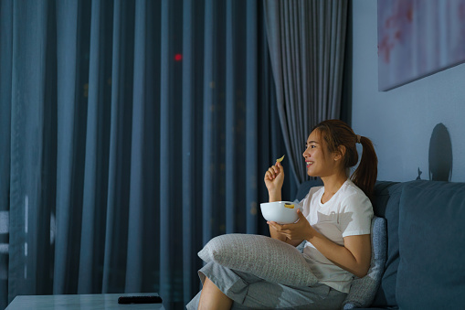 asian woman watching television suspense movie or news looking happy and funny and eating popcorn late night at home living room couch during time of home isolation.