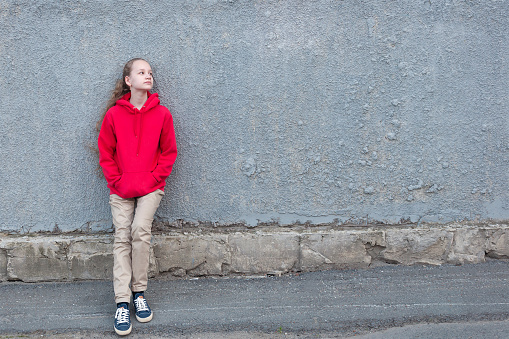 A young blonde girl stands tall in a red hoodie, blue sneakers with white laces, and beige chinos on the asphalt, against a gray concrete wall. Looks away. Summer.