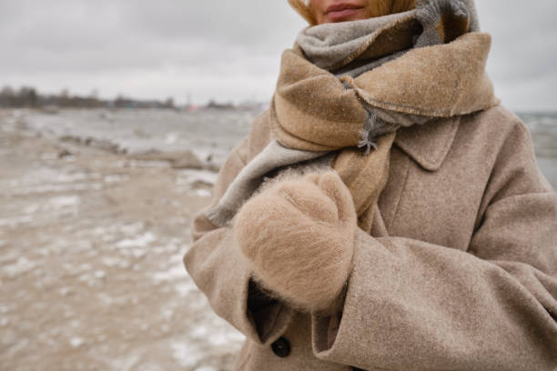 una ragazza elegante si è avvolta in una sciarpa e guanti e cammina lungo il mare o la spiaggia dell'oceano in una giornata invernale. concetto di viaggio. cammina sul mare o sull'oceano. ragazza in viaggio. giornata invernale in riva al mare - wind scarf women people foto e immagini stock