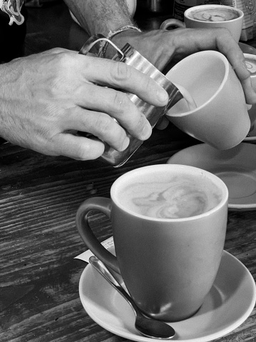 Black and white vertical closeup photo of a Barista, hands only visible, pouring milk from a stainless steel jug into a coffee cup on a wooden bench in a cafe. Ulladulla, NSW