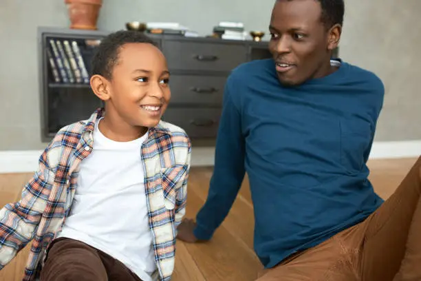 Divorced young African American man in stylish blue jumper spending time with his son on Sunday, both sitting on floor in living-room, talking, smiling, dad looking at him with surprise and admiration