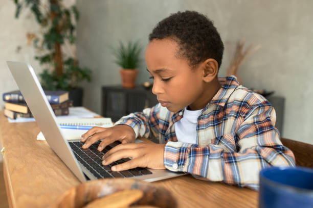 um menino concentrado de 9 anos de etnia africana digitando no teclado do laptop moderno, sentado na mesa de madeira na sala de estar, tendo um olhar sério, fazendo trabalho doméstico. ensino domiciliar. aprendizado distante - 7 10 years - fotografias e filmes do acervo