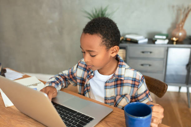 istock Smart brilliant black boy using laptop for studying at home, doing homework, sitting at wooden table against living-room interior, holding blue cup of water or juice in hand, looking at keyboard 1367386109