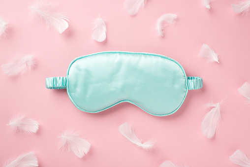 Top view photo of light blue silk sleeping mask and pink feathers on isolated pastel pink background with empty space