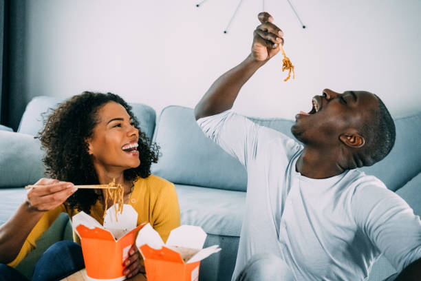 Happy couple eating chinese food at home. Shot of an adorable young couple eating chinese takeout food in carton boxes at home. Young couple sitting on the floor in the living room and having lunch or dinner together. chinese takeout stock pictures, royalty-free photos & images