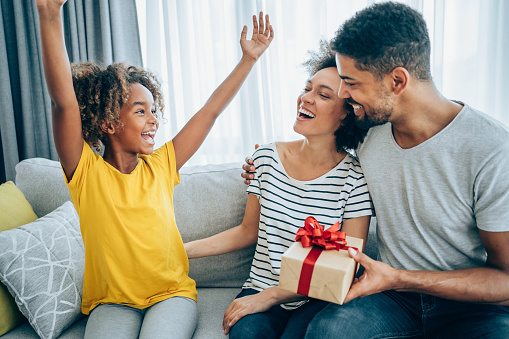 Shot of a loving mother and father giving their daughter a gift. Parents surprise their little girl with present while she is sitting on the sofa in the living room at home.