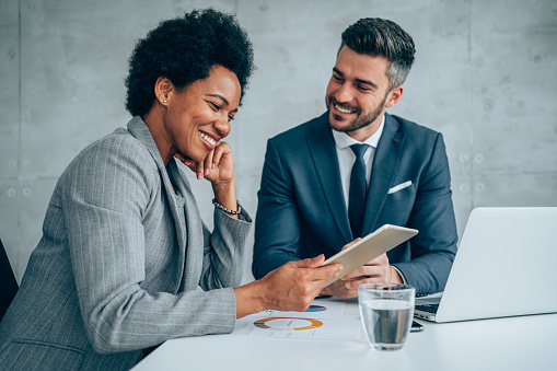 Shot of a two confident business persons sitting on a desk in the office using laptop and digital tablet. Businessman and businesswoman in meeting discussing business strategy. Business coworkers working together in the office.