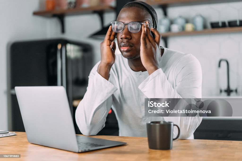 Stressed businessman using laptop at home. Stressed man working from home. Frustrated businessman with head in hands sitting on the table at home. Overworked young afro-american businessman sitting in front of laptop and holding head. Home office concept. African Ethnicity Stock Photo