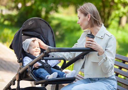 Beautiful Caucasian mother sits on a bench with baby in stroller outdoor