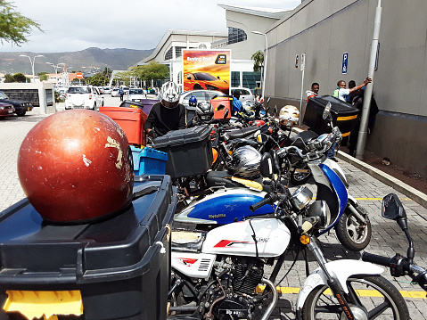 Cape Town, South Africa - January 28, 2022: A large group of motorcycles equipped with boxes outside a shopping mall in the southern suburbs of Cape Town. The riders stand around waiting to be called to deliver fast food. Deliveries of food have increased dramatically in South Africa during the coronavirus pandemic.