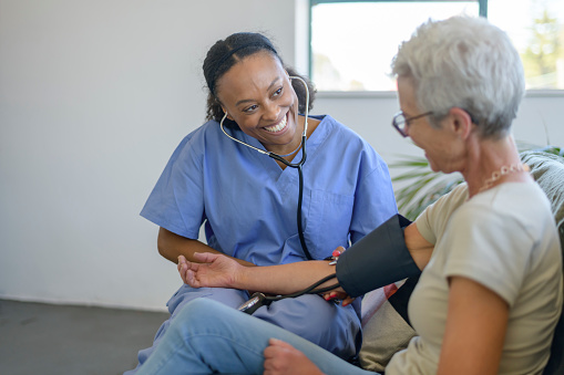 A female mixed race nurse of Hispanic and Latin descent sits on a sofa with her senior patient and checks the aging woman's blood pressure.