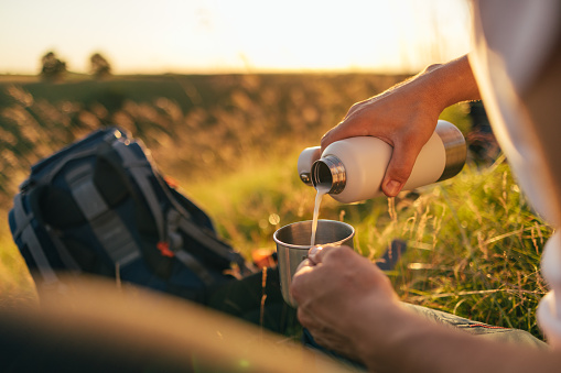 Close up shot of anonymous man camping in nature, holding a cup while sipping milk from a aluminium bottle.