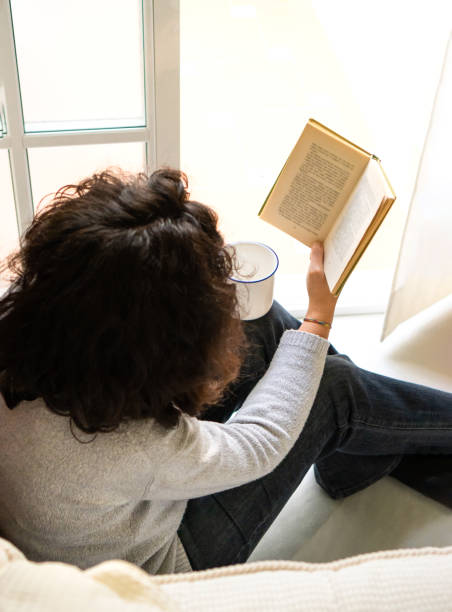 istock Unrecognizable lady drinking coffee and reading book on windowsill 1367417856