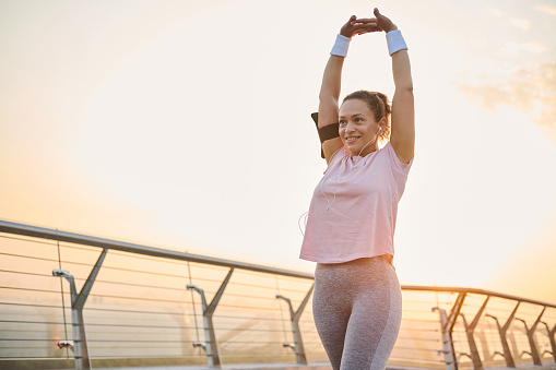 Happy determined female athlete, sportswoman enjoys morning cardio workout, stretches her arms, raising them up, exercises at sunrise. Fitness, health, body care, sport and active lifestyle concept