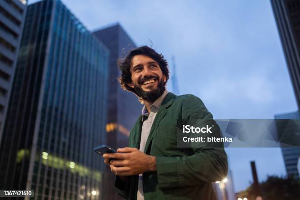 View Of Young Man Using A Smartphone At Night Time With City View Landscape In The Background High Quality Photo Stock Photo - Download Image Now