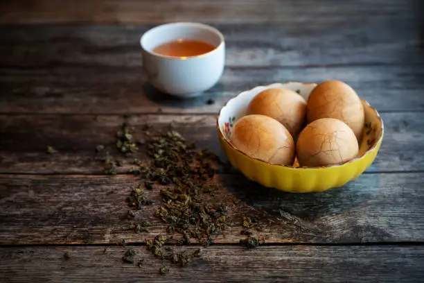 Hard boiled Chinese marble spiced tea eggs on rustic wooden table with tea leaves and cup of Chinese tea in background.