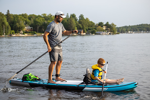 Father and Son Stand-up paddleboarding on quiet lake during a warm sunny summer vacation day. The little boy is sitting on the paddleboard and wearing a life jacket. Lac Saint-Joseph, Fossambault-sur-le-lac, Quebec, Canada.